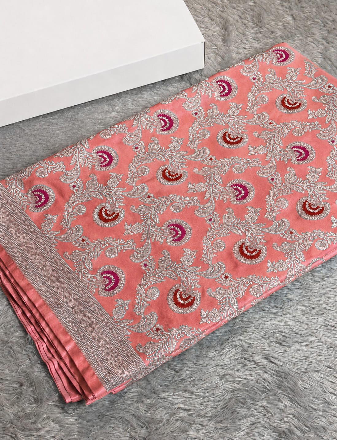 A folded coral pink saree with intricate silver floral embroidery and red and purple paisley motifs, displayed on a gray textured rug next to a white box.