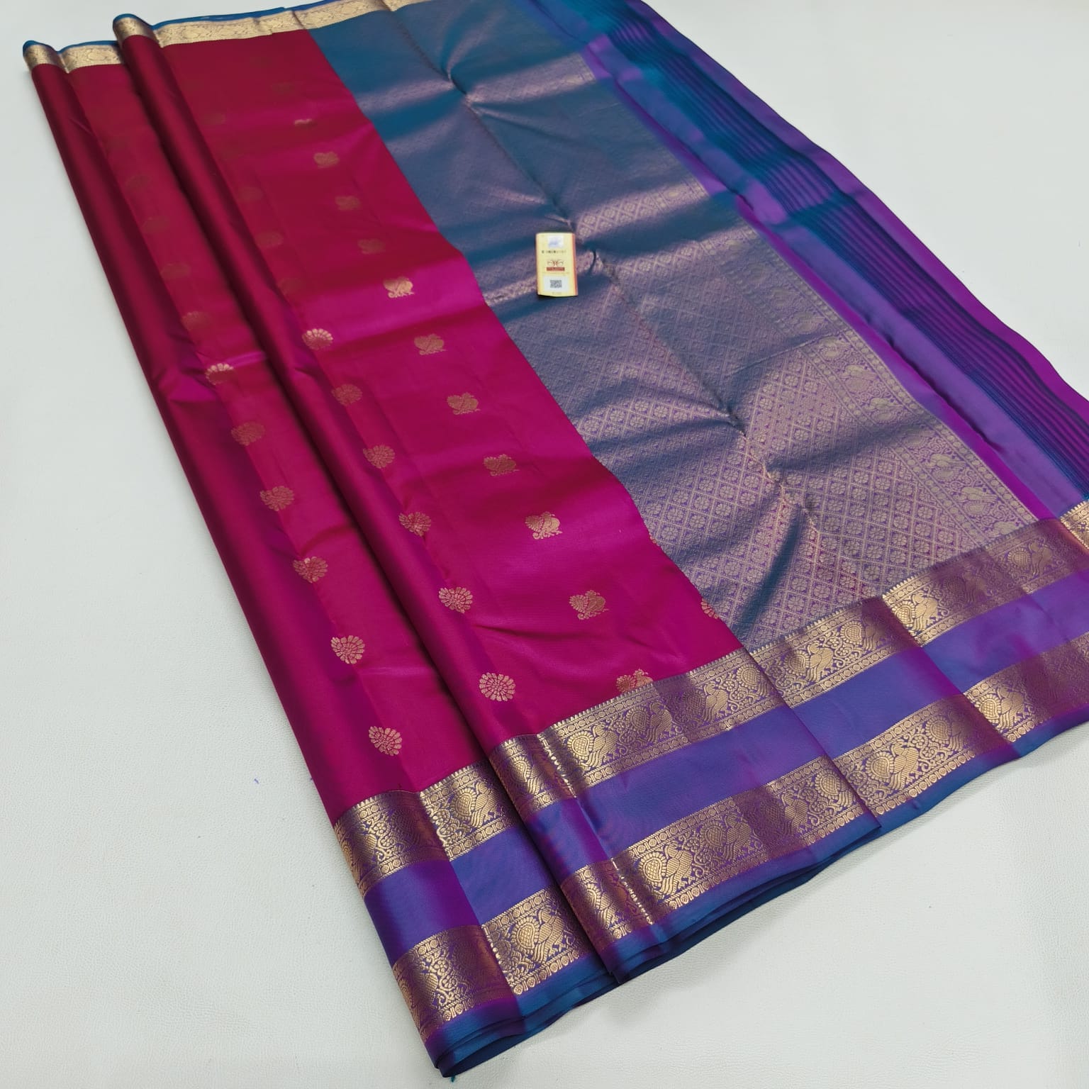 A folded silk saree with a magenta-pink body, a blue and purple border, and intricate gold zari metallic patterns, displayed on a light gray background.