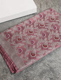 A folded dusty pink saree with intricate silver zari floral embroidery and red and gold paisley motifs, displayed on a gray faux fur surface next to a white box.