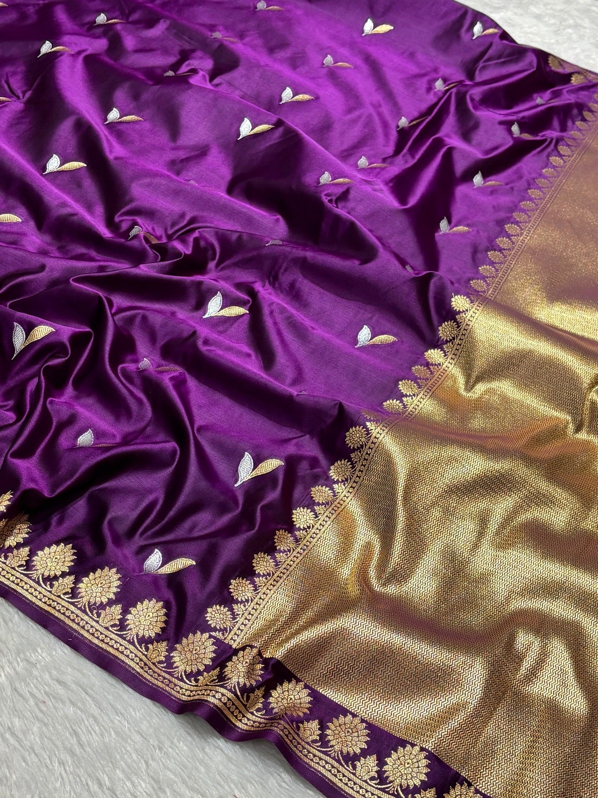 A close-up of a purple silk saree or fabric with small gold and white floral motifs, featuring a wide gold zari border with floral brocade.