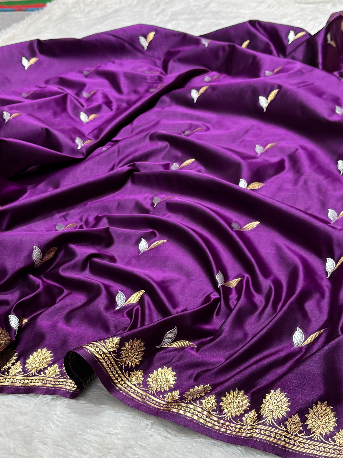 A draped purple silk saree with gold and silver floral and leaf motifs, featuring a decorative gold border, laid out on a white textured surface.