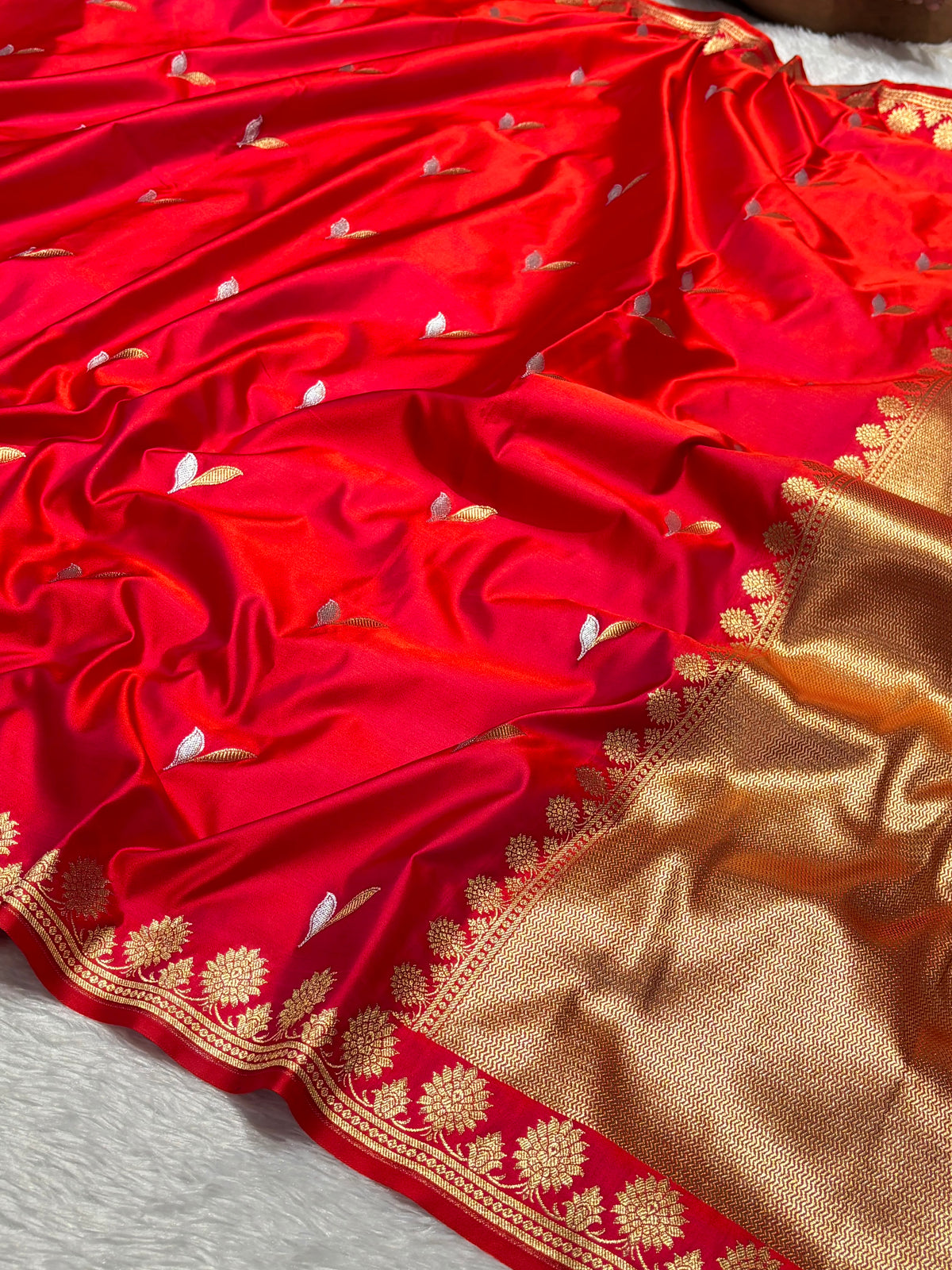 A close-up of a red silk saree with small silver and white floral motifs woven throughout, featuring a wide gold zari border with floral patterns.
