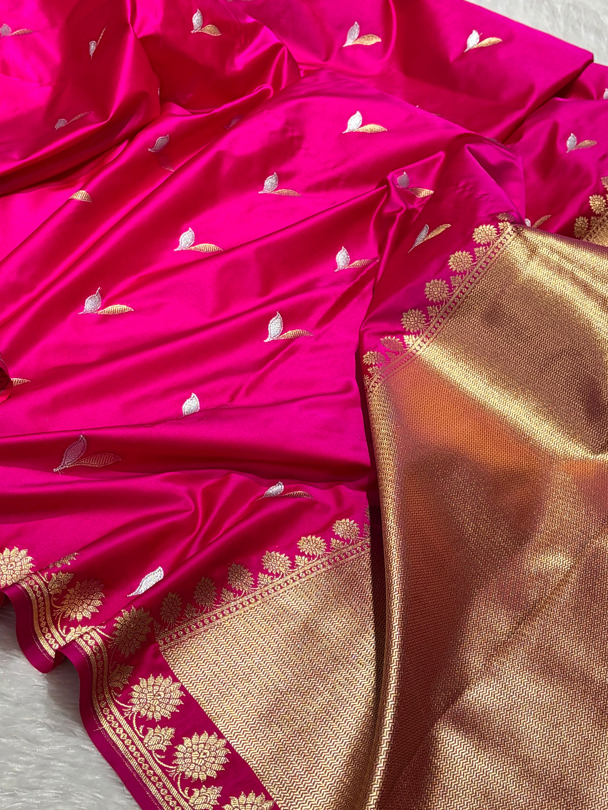A close-up of a bright pink silk saree with small gold and white woven motifs, featuring a wide metallic gold border with floral zari embroidery.