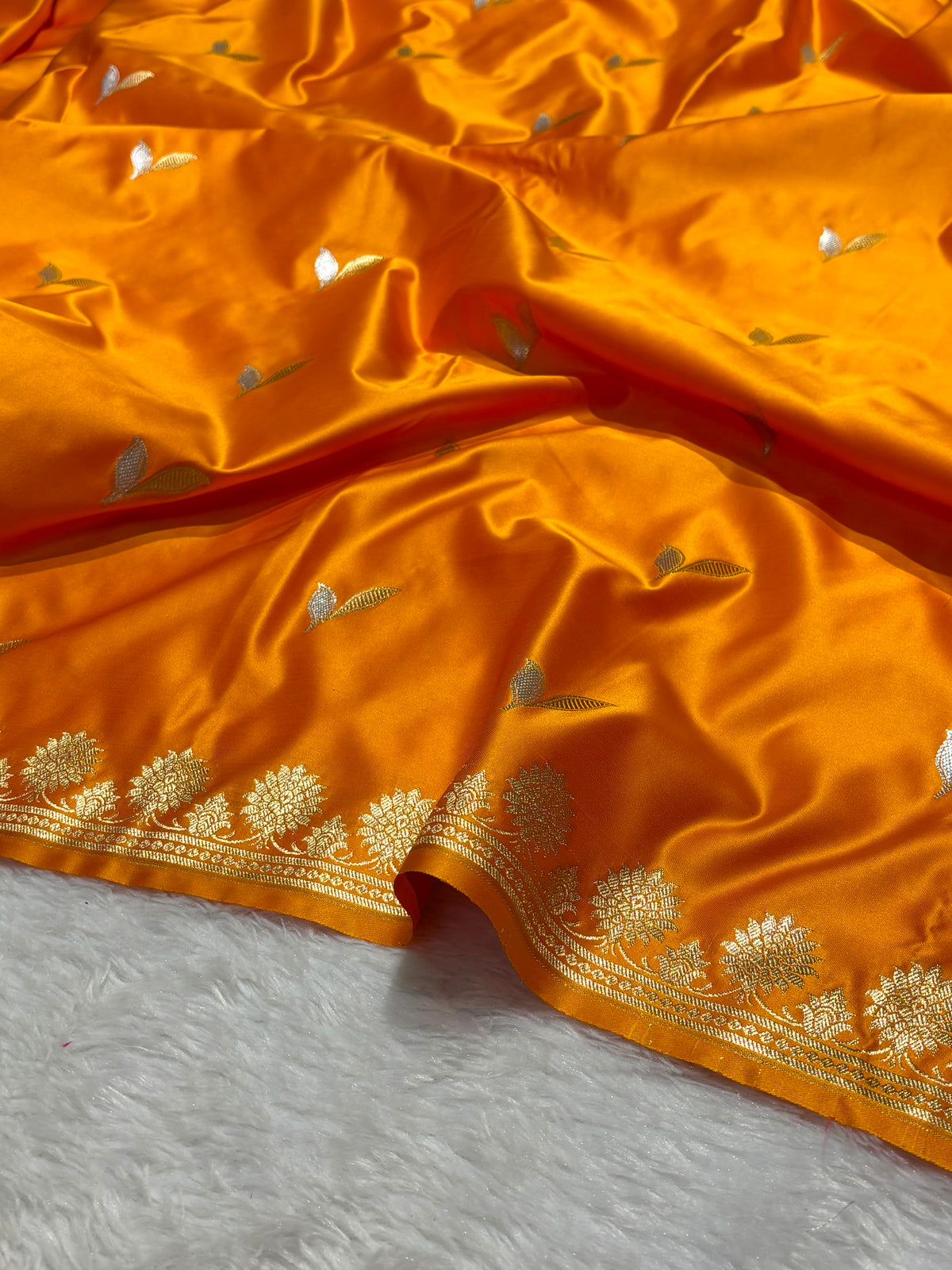 Bright orange silk fabric with metallic gold floral and leaf motifs, draped elegantly on a white textured surface.