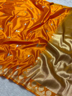 A close-up of an orange silk saree with a gold zari border and small silver motifs woven throughout, draped elegantly on a light gray textured surface.