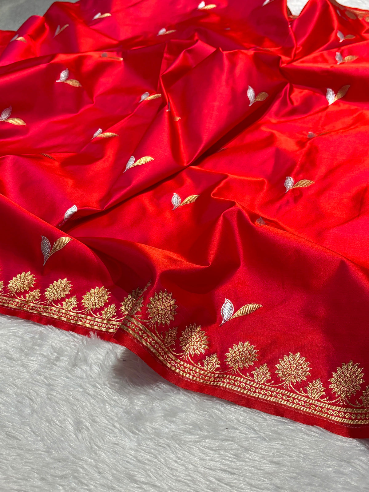 A close-up of a bright red fabric, likely a Banarasi silk saree body, featuring delicate gold and white floral embroidery and a decorative gold border, laid out on a white textured surface.
