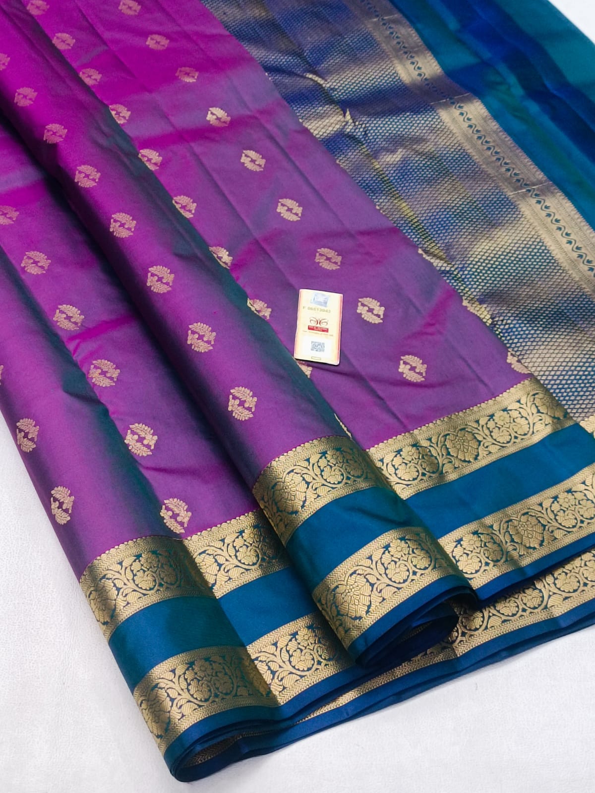 A folded purple and teal silk saree with intricate gold zari floral motifs and borders, displayed on a light gray surface.