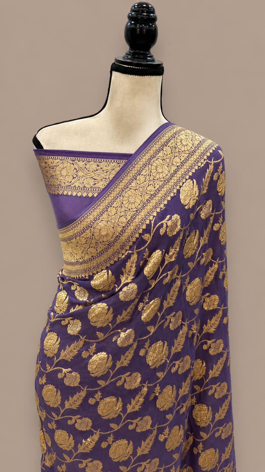 A purple silk saree with intricate gold zari floral patterns and a wide gold border, displayed on a mannequin torso against a solid beige background.