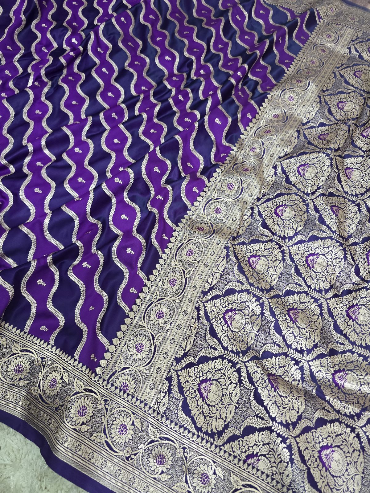 A close-up of a purple and navy blue patterned fabric, likely a silk saree, featuring silver zari woven motifs and borders.