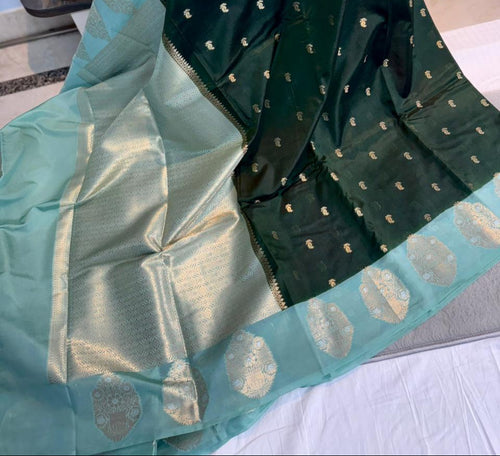 A dark green and light teal green Banarasi silk saree with gold zari woven motifs, held up by a person in a shop setting.