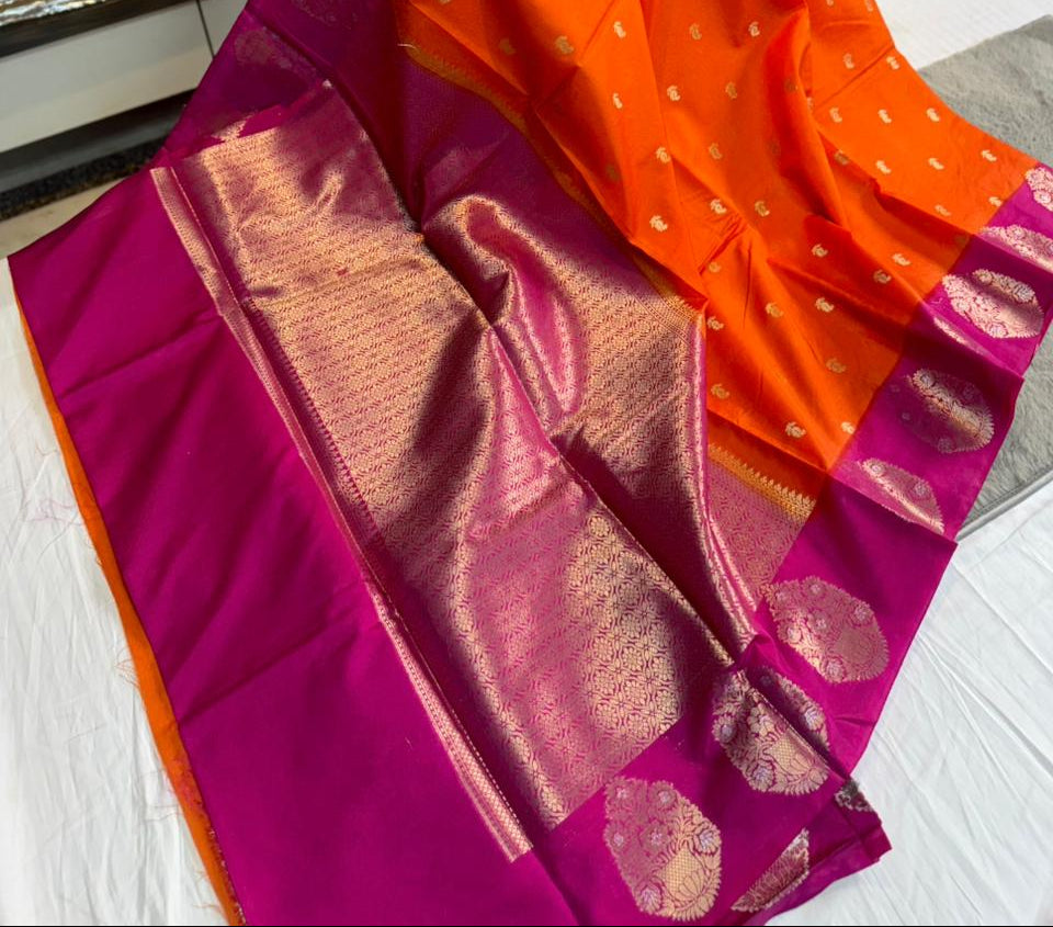 A bright orange and magenta silk saree with gold zari motifs, held up by a person in a shop setting.