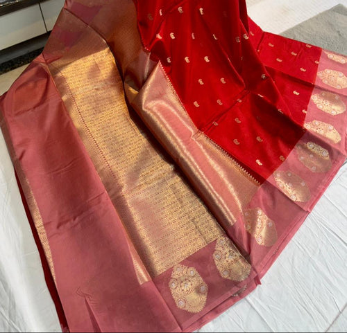 A red and pink silk saree with gold zari brocade work, draped on a white surface with stacks of folded fabric visible in the background.