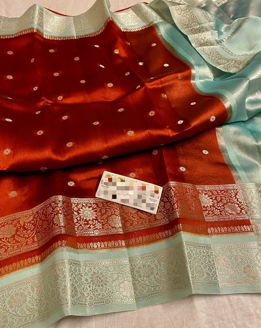 A close-up of a red Banarasi silk saree with silver zari motifs, a light blue dupatta, and a silver border, with a small card placed on the fabric.