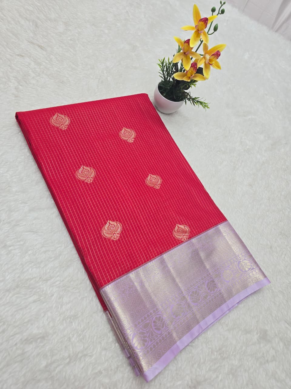Red saree with silver border on a light gray surface with a small plant and yellow flowers.