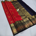 A red and navy blue silk saree with an ornate gold zari border, laid out diagonally on a light gray surface.