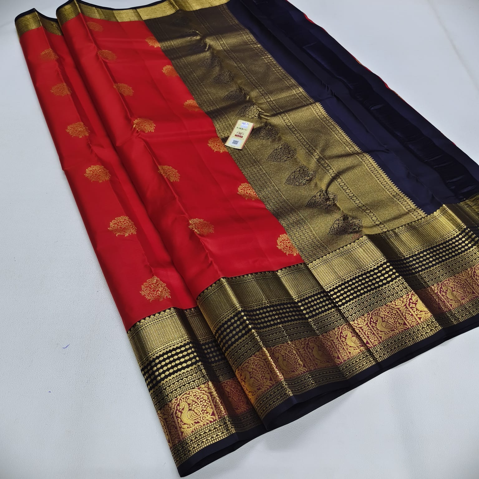A red and navy blue silk saree with an ornate gold zari border, laid out diagonally on a light gray surface.