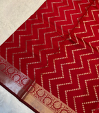 A close-up of a red fabric, likely a saree, featuring a woven beige chevron pattern and a decorative beige border with floral motifs.