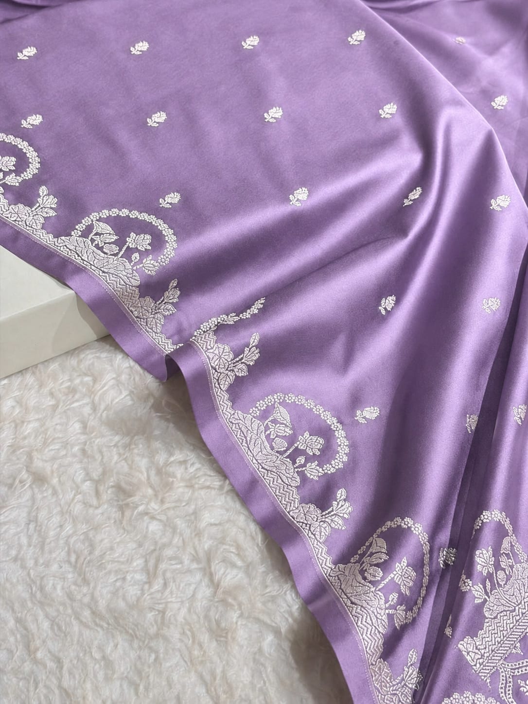 A close-up of a lavender purple fabric, likely a saree or dupatta, featuring intricate silver floral embroidery along the border and scattered small motifs.