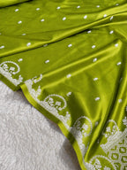 A close-up of an olive green fabric, likely a saree, draped over a white textured blanket. The fabric features small white embroidered dots and a wide white border with detailed floral and geometric patterns.