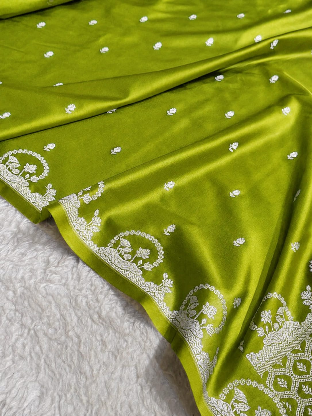 A close-up of an olive green fabric, likely a saree, draped over a white textured blanket. The fabric features small white embroidered dots and a wide white border with detailed floral and geometric patterns.