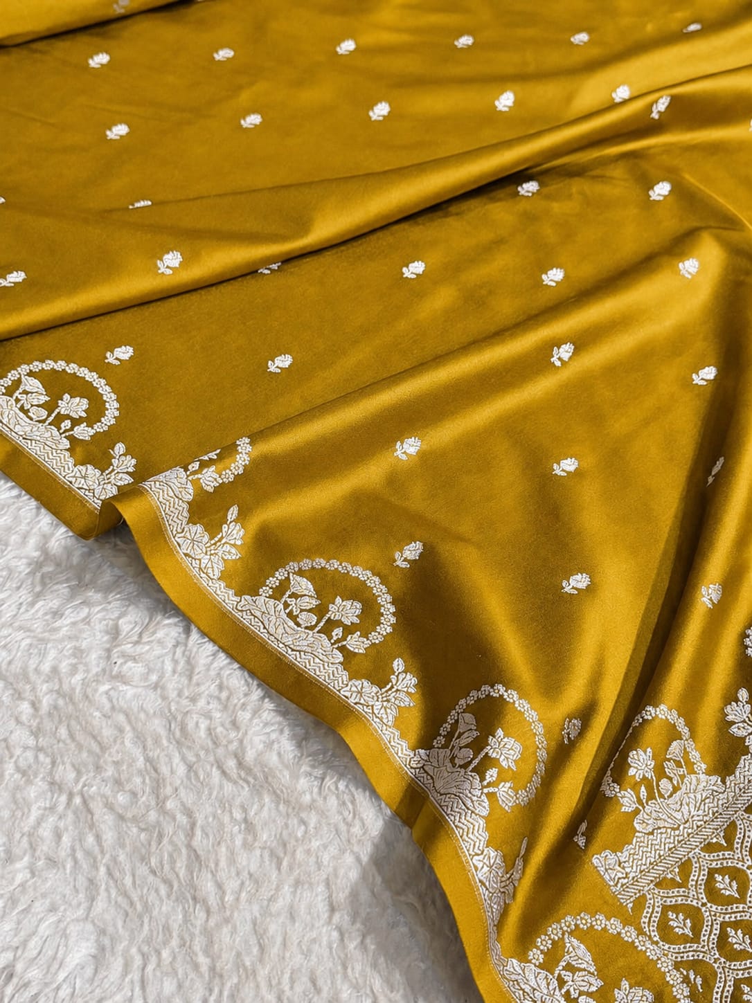 A close-up of a mustard yellow fabric, likely a saree, draped over a white textured surface. The fabric features small white embroidered motifs scattered across it and a prominent white border with detailed floral and geometric embroidery.