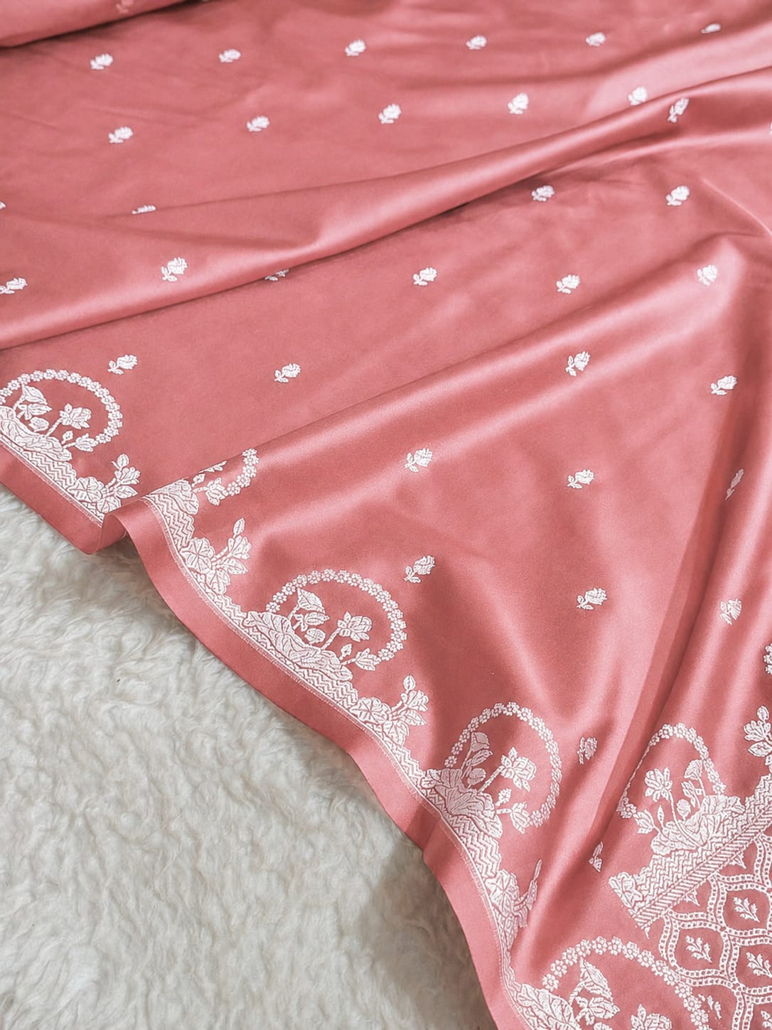 A close-up of a dusty pink fabric, likely a saree, featuring delicate white floral embroidery and a wide white border with traditional motifs, laid over a white textured rug.