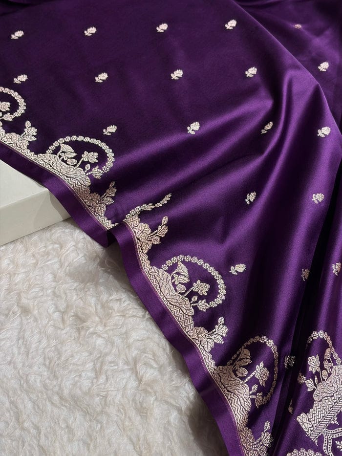 A close-up of a purple fabric, likely a saree or scarf, featuring intricate silver zari embroidery along the border and scattered small motifs, draped over a white textured surface.