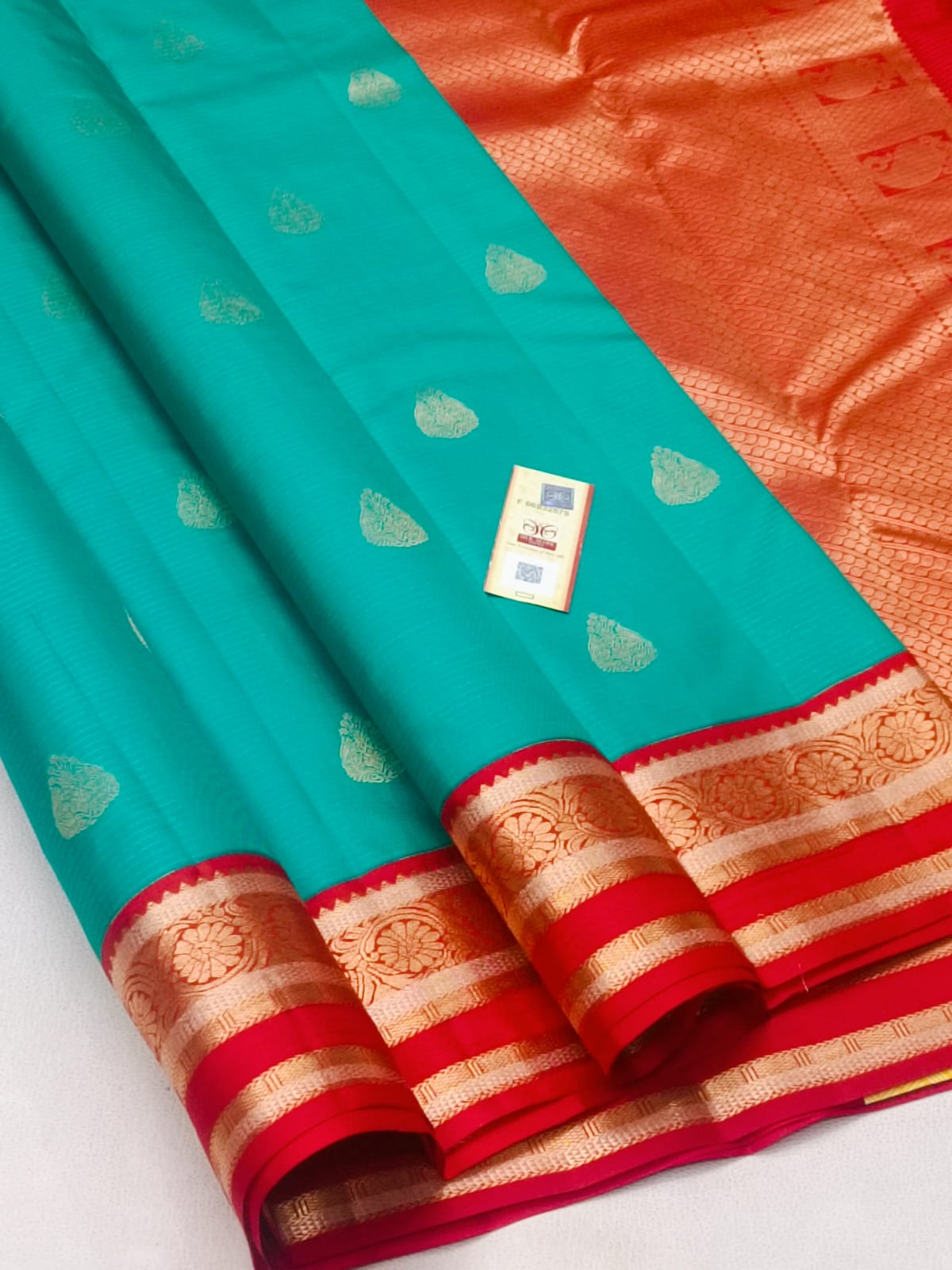 A folded teal silk saree with small metallic motifs, a rich red and gold zari border, and a contrasting orange-red pallu, displayed on a white surface.