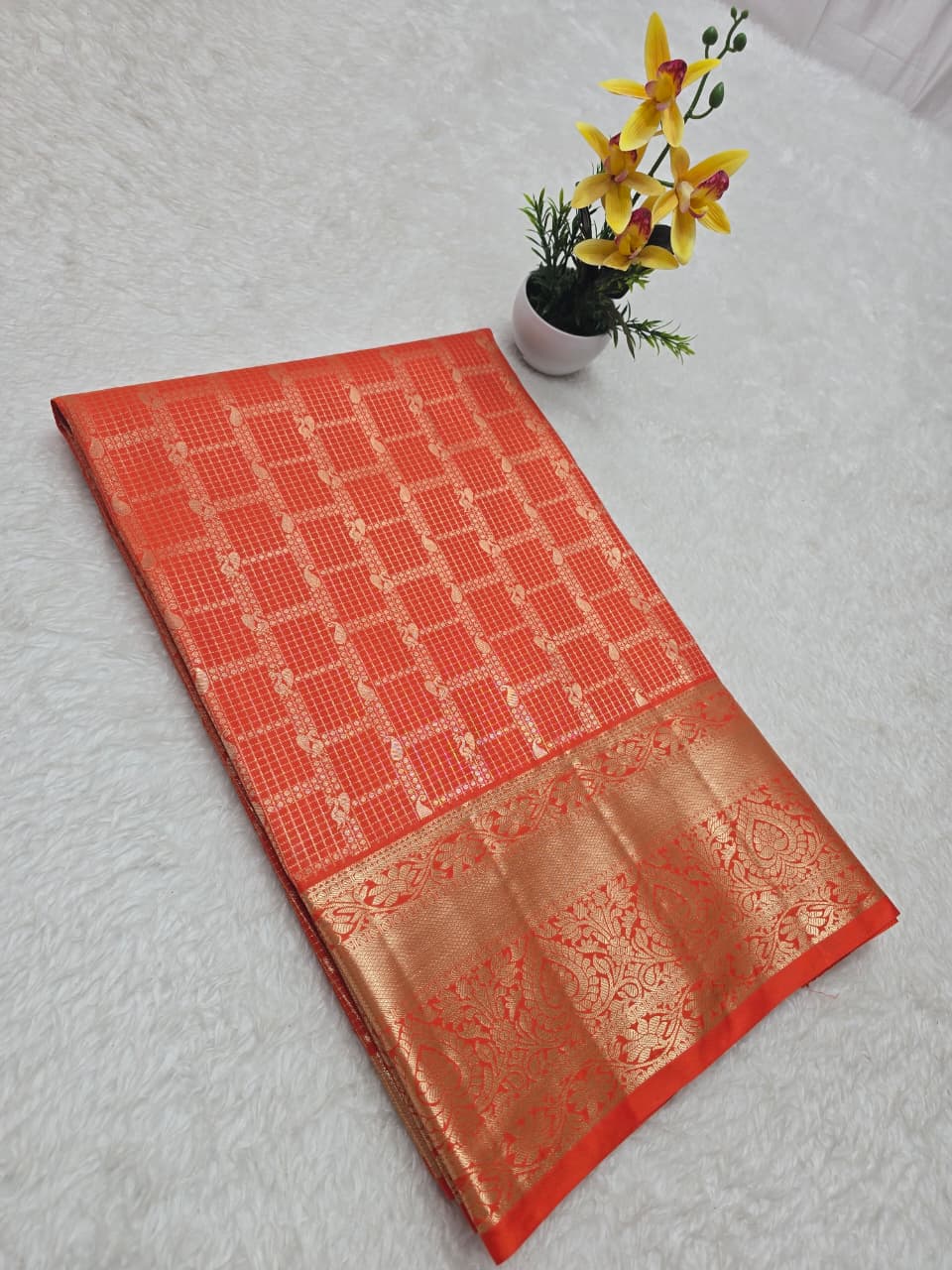 Red and gold saree with a small plant and yellow flowers on a light gray background