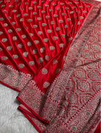 A close-up of a rich red fabric, likely a saree, featuring metallic silver and gold zari embroidery. The fabric is draped to show its texture, pleats, and an ornate border with intricate patterns.