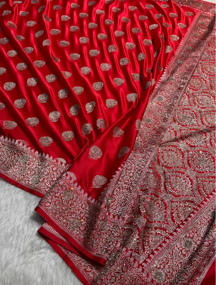 A close-up of a rich red fabric, likely a saree, featuring metallic silver and gold zari embroidery. The fabric is draped to show its texture, pleats, and an ornate border with intricate patterns.