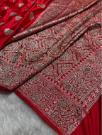 A close-up of a rich red fabric, likely a saree, featuring intricate silver and gold zari embroidery and mirror work. The fabric is draped elegantly to showcase the detailed patterns and sheen of the metallic threadwork.