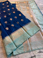 A folded blue and mint green Banarasi silk saree with intricate gold zari embroidery and a wide decorative border, displayed on a marble surface.