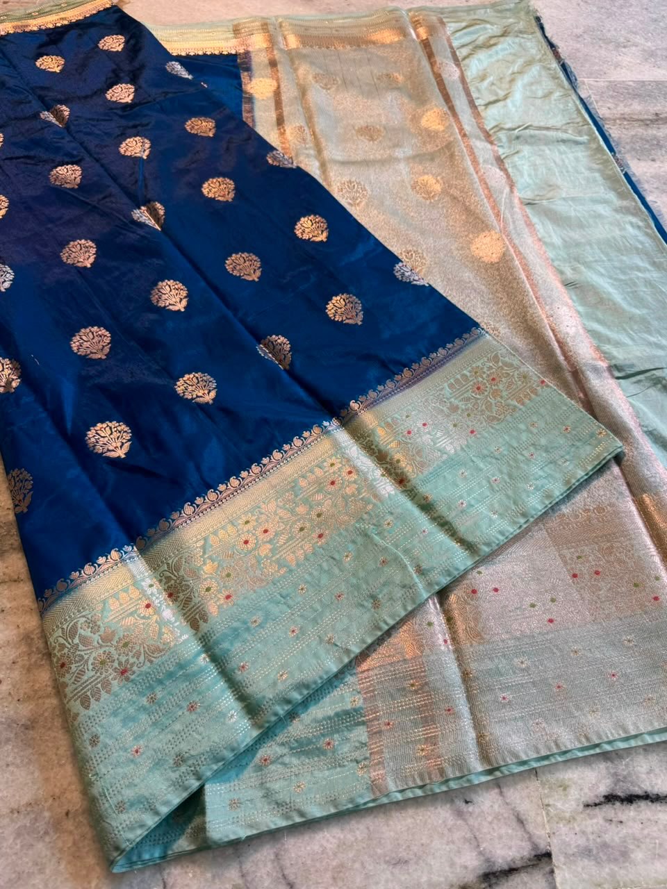 A folded blue and mint green Banarasi silk saree with intricate gold zari embroidery and a wide decorative border, displayed on a marble surface.