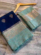 Two folded silk sarees displayed on wooden hangers over a gray marble floor. The saree on the left is navy blue with a mint green border featuring gold and red zari embroidery.