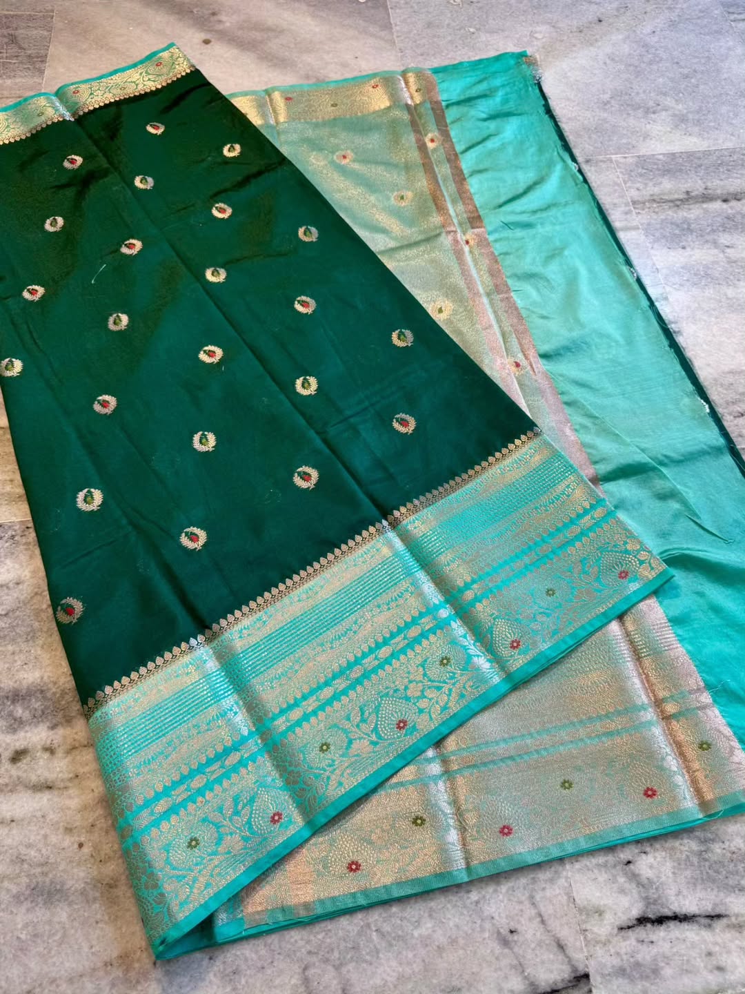 A dark green silk saree with a wide turquoise and gold zari border, laid out diagonally on a gray stone floor.