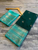 A folded dark green and turquoise saree fabric with intricate gold zari embroidery, displayed on a gray stone floor next to a wooden hanger.