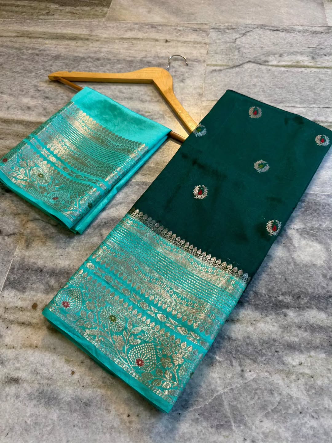 A folded dark green and turquoise saree fabric with intricate gold zari embroidery, displayed on a gray stone floor next to a wooden hanger.