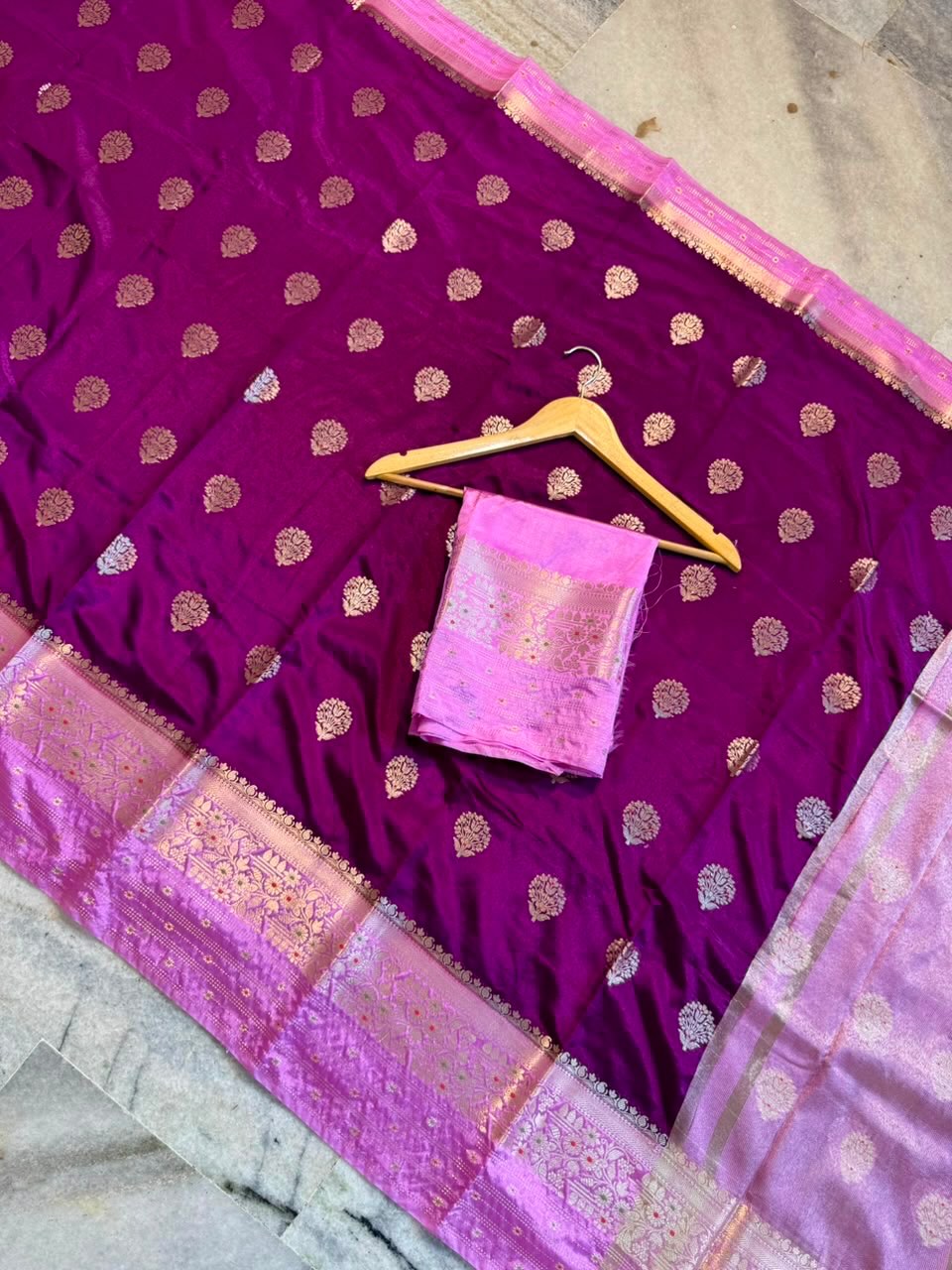 A deep magenta purple Banarasi silk saree with gold zari motifs and a pink border, displayed with a matching pink blouse piece on a wooden hanger.