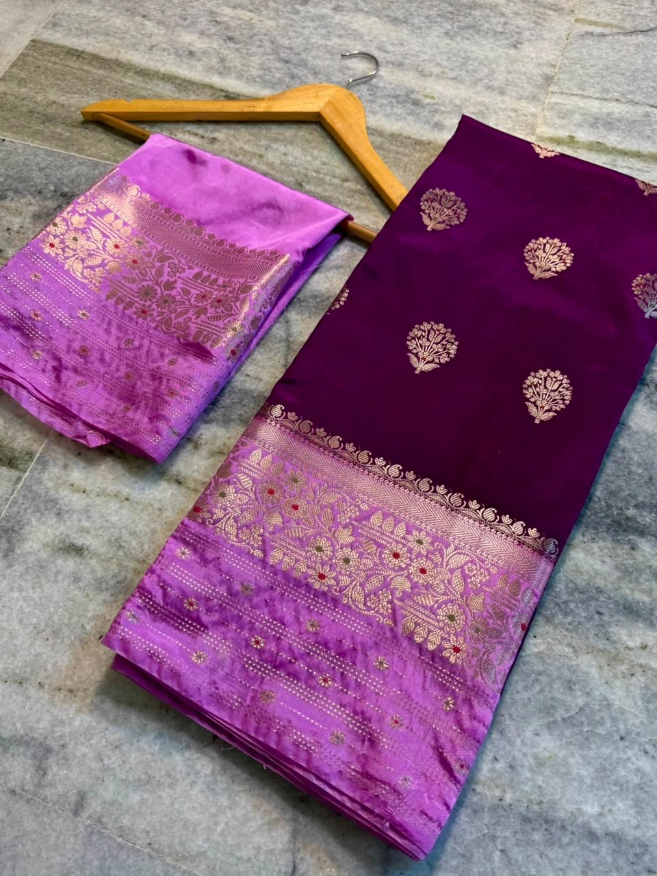 A folded purple and magenta silk saree with intricate gold zari embroidery, displayed on a wooden hanger over a gray stone floor.