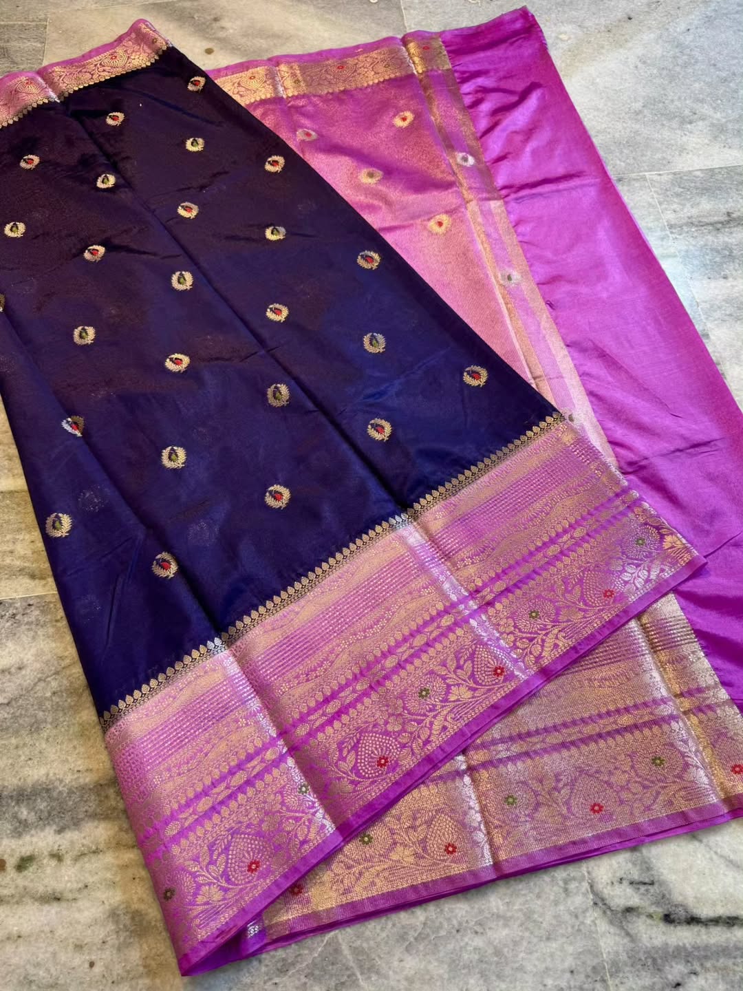 A folded purple and magenta Banarasi silk saree with intricate gold zari brocade borders and small metallic motifs, laid flat on a gray marble floor.