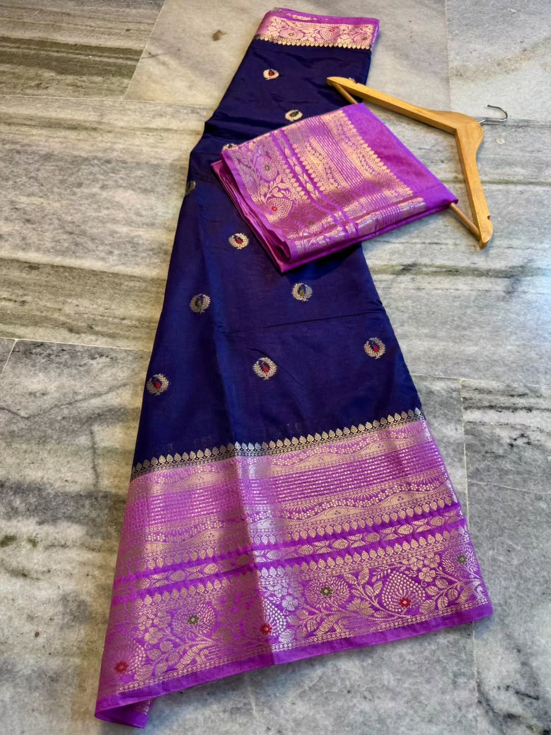 A dark purple Banarasi silk saree with a vibrant magenta and gold zari border, laid out on a gray marble floor next to a wooden hanger.