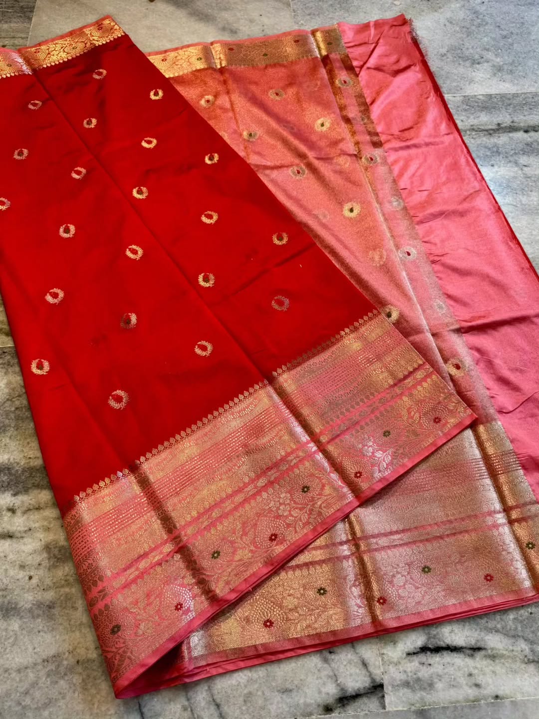 A folded red and pink silk saree with intricate gold zari brocade borders and motifs, displayed on a gray marble floor.