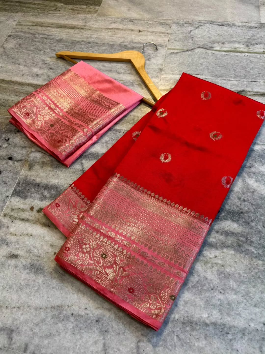 A folded red and pink Banarasi silk saree with intricate gold zari brocade work, displayed on a gray marble floor next to a wooden hanger.