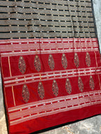 A close-up of a traditional Indian saree fabric, featuring a black and gold striped body with small metallic dots, a wide red border with gold zari embroidery, and ornate gold paisley motifs.