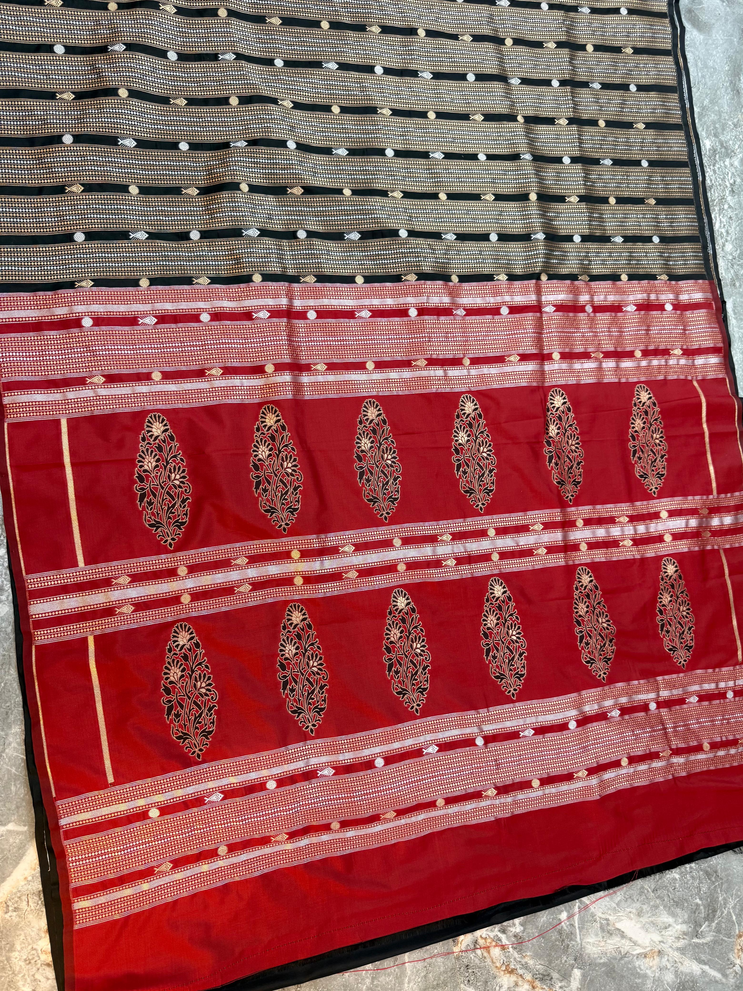 A close-up of a traditional Indian saree fabric, featuring a black and gold striped body with small metallic dots, a wide red border with gold zari embroidery, and ornate gold paisley motifs.