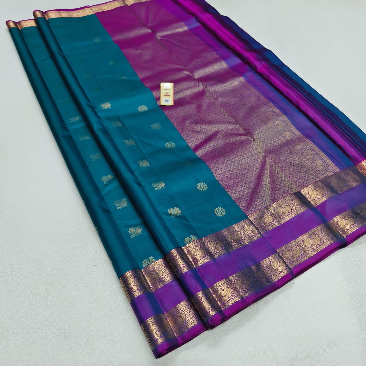 A teal blue and purple silk saree with intricate gold zari woven patterns and borders, displayed diagonally on a light gray background.