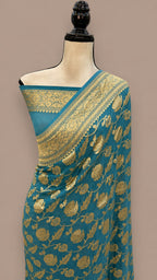 A teal blue saree with intricate gold floral and paisley zari work, draped on a cream-colored mannequin torso against a solid taupe background.