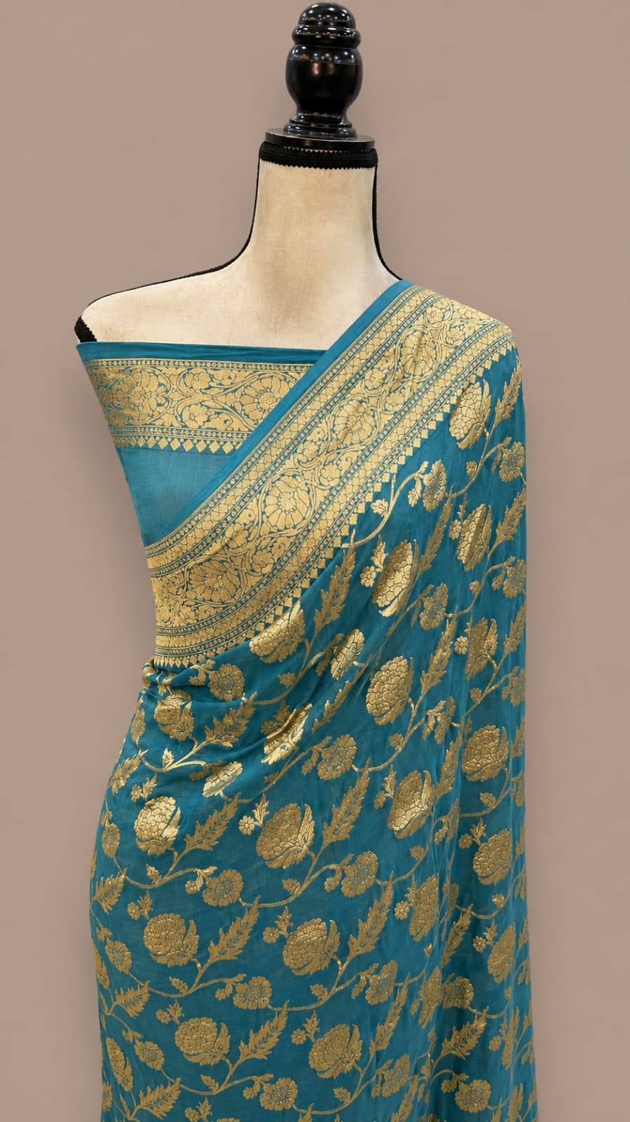 A teal blue saree with intricate gold floral and paisley zari work, draped on a cream-colored mannequin torso against a solid taupe background.