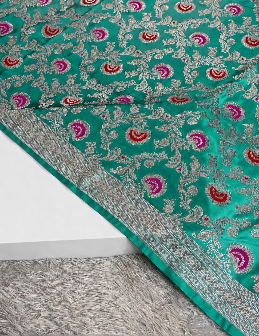 A close-up of a teal green silk saree with intricate silver zari floral patterns and pink and red paisley motifs, draped over a white block with a gray shag rug underneath.